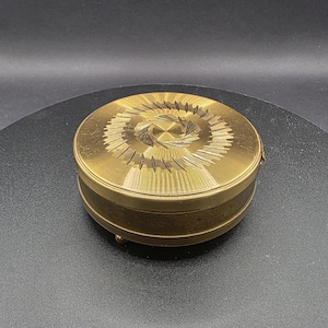 May include: A small, round, antique brass box with an etched design on the lid. The lid has a central floral motif surrounded by radiating lines. The box has a hinged lid and small feet.
