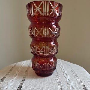 May include: A ruby red cut-glass vase with a distinctive stacked design. The vase has intricate star and geometric patterns, adding elegance. This decorative piece is ideal for displaying flowers or as a standalone art object. It is approximately 20 cm tall.