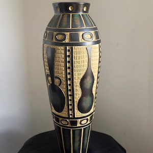 May include: A tall, decorative vase in black and gold. The vase has a textured, carved design with oval and vertical shapes, and two stylized, dark-coloured figures. The vase is displayed on a black surface.
