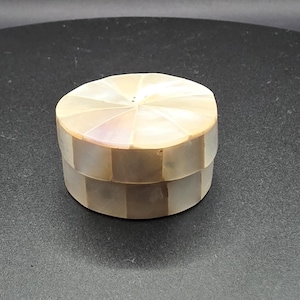 May include: A small, round decorative box with a lid. The box is made of iridescent, pearlescent material, with a geometric pattern on the lid. The sides of the box are a light brown colour. The box is likely used for storing small items.