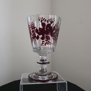 May include: A clear glass goblet featuring a deep red floral and leaf pattern etched around the bowl. The stem and circular base are accented with matching deep red rings, providing a cohesive look to this decorative drinking vessel.