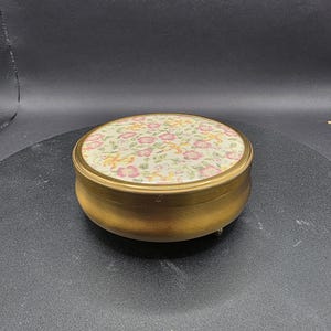 May include: A small, round, gold-toned trinket box with a floral design on the lid. The lid has a cream background with pink and yellow flowers and green leaves. The box has a simple, elegant design.