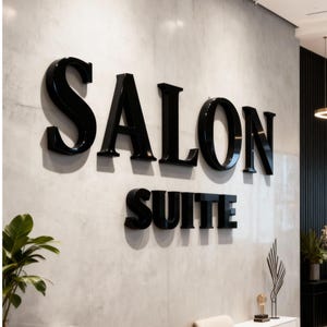 May include: Interior shot of a modern salon. The words "SALON SUITE" are displayed in large, glossy black letters on a textured gray wall. A reception desk with a white countertop and black cabinets is in the foreground. A black salon chair is visible.