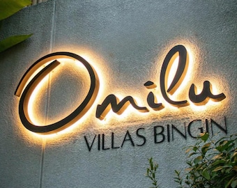 May include: A wall-mounted sign featuring the word Omilu in large, dark, backlit script lettering, with the text VILLAS BINGIN in smaller, dark, non-illuminated capital letters positioned directly underneath on a textured gray wall.