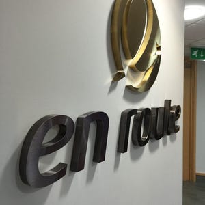 May include: A wall-mounted sign with the words "en route" in a dark metallic finish. Above the text is a gold-colored, abstract logo. The sign is mounted on a light gray wall, with a hallway visible in the background.