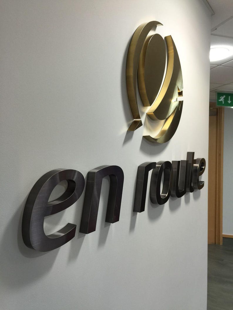 May include: A wall-mounted sign with the words "en route" in a dark metallic finish. Above the text is a gold-colored, abstract logo. The sign is mounted on a light gray wall, with a hallway visible in the background.