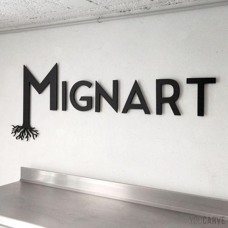 May include: Black wooden sign spelling out "MIGNART" on a white wall. The letter "M" has a root design extending from the bottom. The sign is above a stainless steel surface.