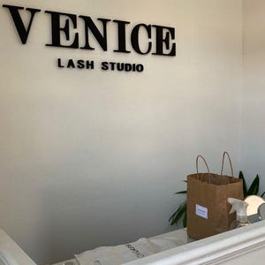 May include: The interior of a lash studio with the words "VENICE LASH STUDIO" in black lettering on a white wall. A brown paper bag and a spray bottle sit on a white counter.
