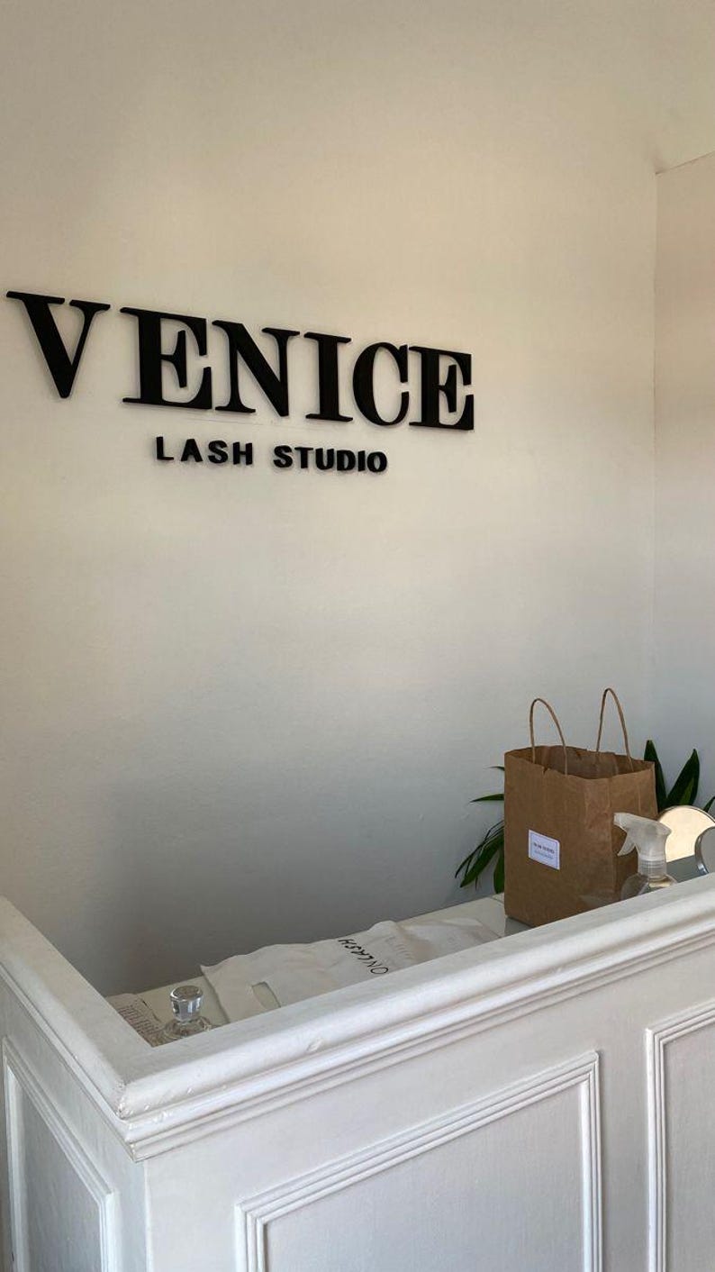 May include: The interior of a lash studio with the words "VENICE LASH STUDIO" in black lettering on a white wall. A brown paper bag and a spray bottle sit on a white counter.
