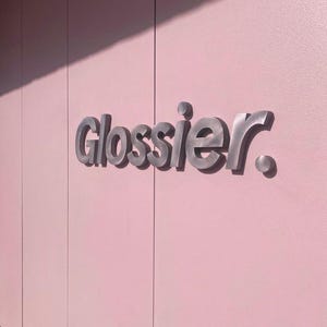 May include: The word "Glossier" in silver, three-dimensional lettering is mounted on a light pink wall. The wall is divided into vertical panels with subtle shadows. The overall aesthetic is clean and modern.