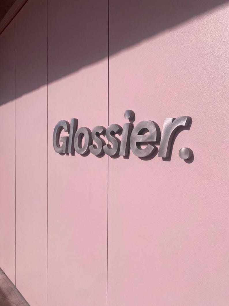 May include: The word "Glossier" in silver, three-dimensional lettering is mounted on a light pink wall. The wall is divided into vertical panels with subtle shadows. The overall aesthetic is clean and modern.