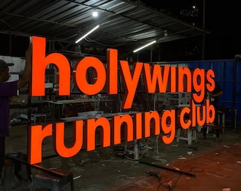 May include: Large, illuminated orange sign that reads "holywings running club." The sign is made of individual, three-dimensional letters. The sign is outdoors, likely at night, with a person working on the sign.