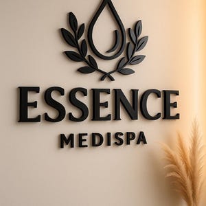 May include: Black 3D sign on a beige wall. The sign features the word "ESSENCE" above "MEDISPA". A black water droplet design with leaves is above the text. A vase of dried plants is in the lower right corner.