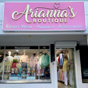 May include: Exterior shot of Arianna's Boutique, a resort wear, apparel, and accessories store. The store's sign is pink with gold lettering. Mannequins in the window display clothing, including shirts, dresses, and hats. The store's entrance is open.