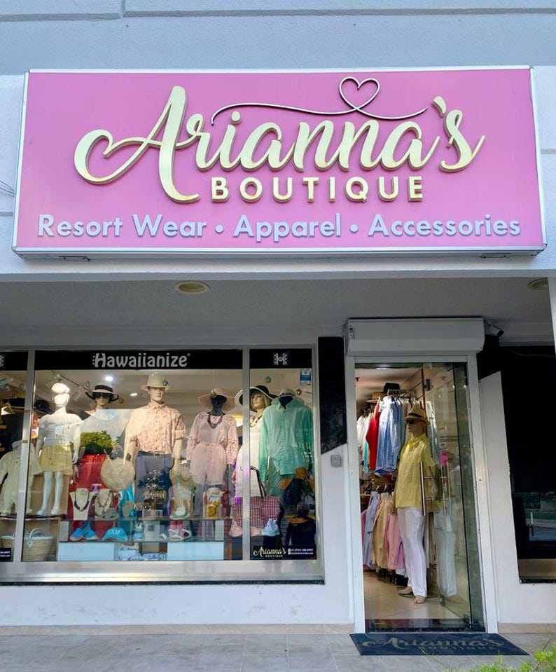 May include: Exterior shot of Arianna's Boutique, a resort wear, apparel, and accessories store. The store's sign is pink with gold lettering. Mannequins in the window display clothing, including shirts, dresses, and hats. The store's entrance is open.