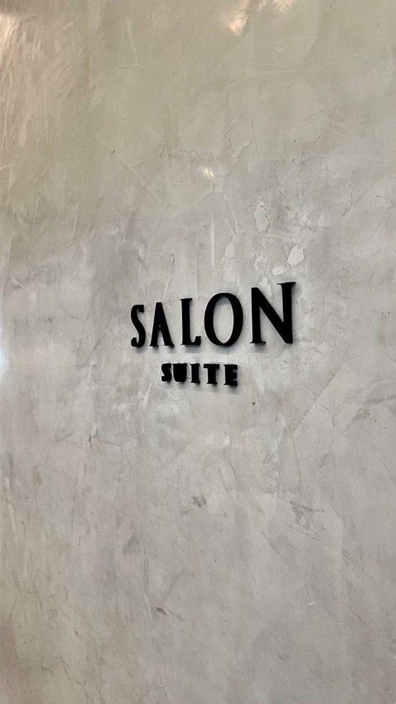 May include: Black, raised lettering on a textured, light gray wall reads "SALON SUITE." The word "SALON" is above "SUITE." The wall has a mottled appearance, with variations in tone and texture.