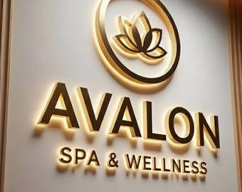 May include: A gold-colored illuminated sign for "Avalon Spa & Wellness" on a cream-colored wall. The sign features a lotus flower emblem within a circle, and the text is in a bold, sans-serif font. The overall design is elegant and modern.