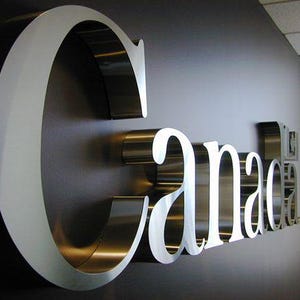 May include: A metallic sign spelling "Canada" is mounted on a dark wall. The letters are silver with a white outline, creating a three-dimensional effect. The sign is a decorative element.