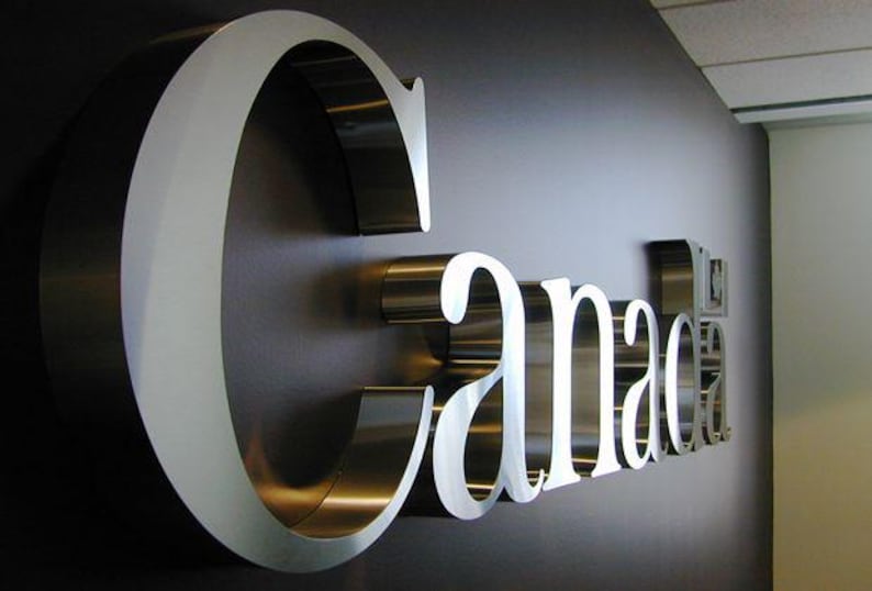 May include: A metallic sign spelling "Canada" is mounted on a dark wall. The letters are silver with a white outline, creating a three-dimensional effect. The sign is a decorative element.