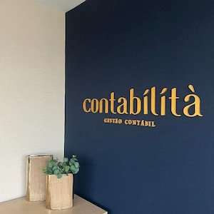 May include: A navy blue wall with gold lettering that reads "contabilit&agrave;" and "gest&atilde;o cont&aacute;bil". A light beige wall is adjacent to the navy blue wall. Two decorative vases with greenery sit on a light wood surface.