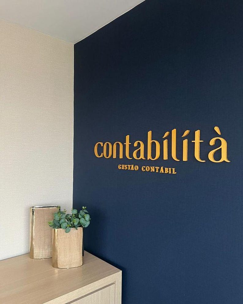 May include: A navy blue wall with gold lettering that reads "contabilit&agrave;" and "gest&atilde;o cont&aacute;bil". A light beige wall is adjacent to the navy blue wall. Two decorative vases with greenery sit on a light wood surface.
