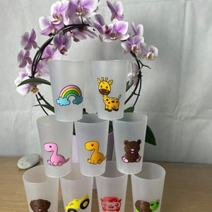 Può includere: Una collezione di bicchieri di plastica smerigliata, ognuno decorato con un colorato disegno a cartone animato. I disegni includono un arcobaleno, una giraffa, un dinosauro, un orso, un cucciolo, un camion, un'auto e una tartaruga. I bicchieri sono impilati, con fiori sullo sfondo.