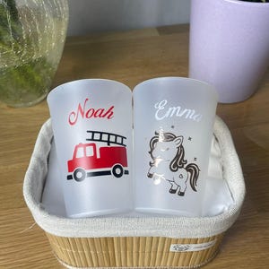 Może przedstawiać: Dwa szklanki z matowego szkła z personalizowanymi wzorami. Jeden ma czerwoną straż pożarną i imię "Noah" w czerwonym skrypcie. Drugi ma grafikę jednorożca i imię "Emma" w srebrnym skrypcie. Szklanki znajdują się w wiklinowym koszu.