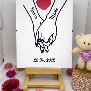 Puede incluir: Un letrero blanco sobre un caballete de madera presenta un dibujo de dos manos tomadas de la mano, con los nombres "Simon" y "Marie" y un corazón rojo. La fecha "23 Fev 2012" está debajo. También hay flores de papel y un osito de peluche.