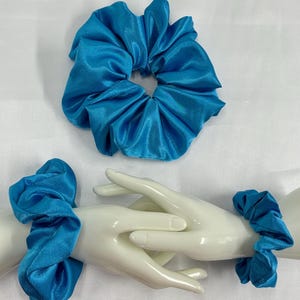 May include: Two turquoise satin scrunchies. One scrunchie is displayed on its own, while the other two are worn on the wrists of a white mannequin hand. The scrunchies are made of a soft, shiny fabric.