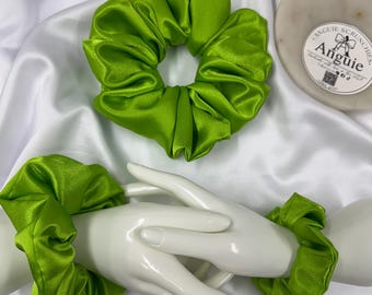 May include: Two bright green satin scrunchies. One scrunchie is displayed on a white surface, and the other two are worn on the wrists of a white mannequin hand. The scrunchies are made of a shiny, smooth fabric.