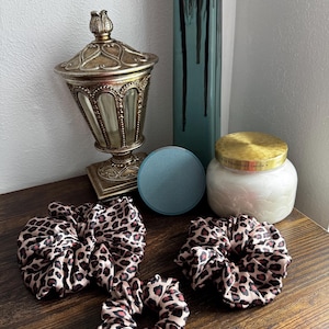 May include: A collection of leopard print scrunchies in various sizes, displayed on a wooden surface. The scrunchies feature a brown and black spotted pattern. Decorative items, including a gold-toned jar, a teal vase, and a candle, are in the background.
