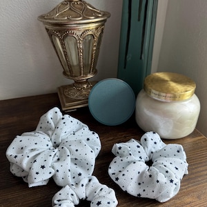 May include: Three white scrunchies with a navy blue star pattern are arranged on a wooden surface. A decorative gold and glass container, a teal coaster, and a white candle with a gold lid are also present.