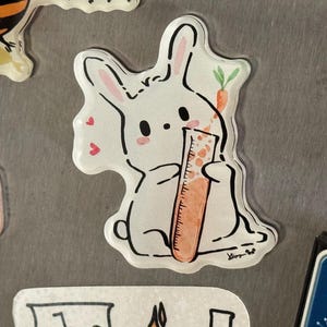 May include: A white bunny sticker with black outlines, pink ears, and a carrot. The bunny is holding a graduated cylinder. Small pink hearts are scattered around the bunny. The sticker is on a metallic surface.