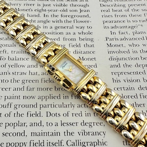 May include: A gold Citizen Elegance watch with a rectangular face and a mother-of-pearl dial. The watch has a linked bracelet design. The watch is displayed on a printed page.