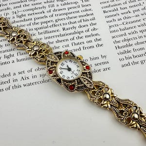May include: A vintage gold-toned watch with an ornate design. The watch face is white with black numerals and hands, accented by red jewels. The band features intricate floral and geometric patterns, creating a classic, elegant aesthetic.