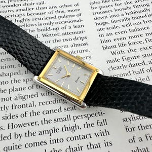 May include: A vintage Citizen Quartz watch with a rectangular gold-toned case and a silver dial. The watch features gold hands and markers, and is set on a black textured leather strap. The word "Seven" is printed on the dial.