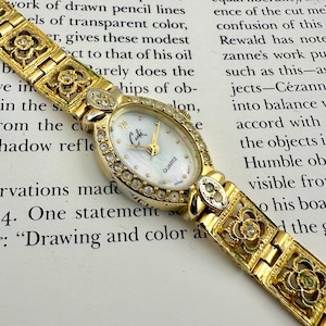 May include: A gold-toned Cache quartz watch with an oval face and a mother-of-pearl dial. The watch features a bezel and band adorned with clear crystals and decorative floral cutouts. The watch band is approximately 7 inches long.
