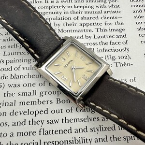 Puede incluir: Un reloj de pulsera Bulova con caja cuadrada de tono plateado y esfera color crema con marcadores de hora y manecillas plateadas. El reloj está sujeto a una correa de cuero marrón oscuro con costuras blancas visibles en los bordes.