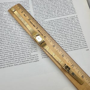 K&ouml;nnte beinhalten: Eine goldfarbene Armbanduhr mit rechteckigem Zifferblatt und Mesh-Armband liegt auf einem Holzlineal. Die Uhr befindet sich in der N&auml;he der Mitte des Bildes, wobei die Markierungen des Lineals sichtbar sind.