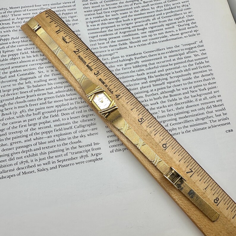K&ouml;nnte beinhalten: Eine goldfarbene Armbanduhr mit rechteckigem Zifferblatt und Mesh-Armband liegt auf einem Holzlineal. Die Uhr befindet sich in der N&auml;he der Mitte des Bildes, wobei die Markierungen des Lineals sichtbar sind.