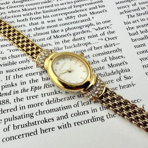 Könnte beinhalten: Eine goldfarbene Elgin Armbanduhr mit einem weißen, ovalen Zifferblatt und Kristallakzenten. Die Uhr hat ein gewebtes gold- und braunes Armband. Die Uhr liegt vor einem Hintergrund mit gedrucktem Text.