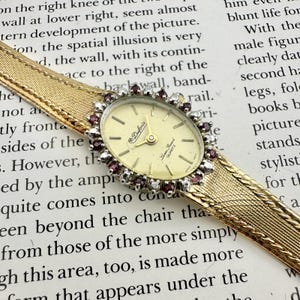 May include: A vintage Dufonte quartz watch with a gold-tone band and a cream-colored oval face. The watch face is surrounded by a halo of small, red gemstones. The band has a woven design.