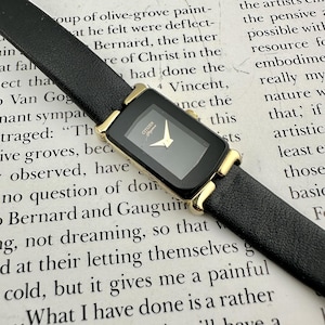 May include: A black Citizen watch with a rectangular face and gold-tone accents. The watch has a black leather band. The watch face features gold hands and the brand name. The watch is set against a background of printed text.