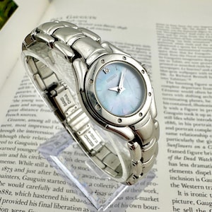May include: A silver-tone Seiko wristwatch featuring a round case with a light blue mother-of-pearl dial, a single diamond marker at the twelve o'clock position, silver hands, and a polished stainless steel link bracelet with a secure clasp.