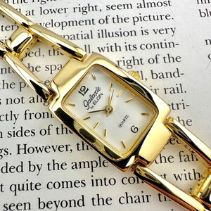 May include: A gold-toned Galaxie by Elgin quartz watch with a rectangular face and a white dial. The watch features gold hands and hour markers, and a gold link bracelet. The watch face displays the number 12 and the number 6.