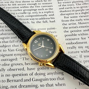 Pode incluir: Um relógio analógico dourado com mostrador preto e pulseira de couro preta. O relógio tem um mostrador redondo com ponteiros e marcadores de hora dourados. O mostrador do relógio exibe a marca "Tenor Ingrido" e a palavra "Quartz".