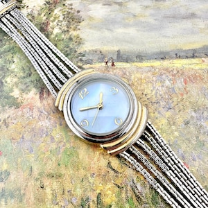 May include: A vintage wristwatch with a light blue face and gold-tone hands and accents. The watch has a silver-tone band with multiple strands. The watch face features the numbers 12, 3, and 6. The watch is set against a colorful background.