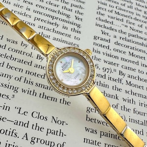 May include: A gold-coloured Anne Klein II wristwatch with a mother-of-pearl face and a bezel set with small, clear crystals. The watch features a rectangular link band and the text "2000" is visible on the watch face.