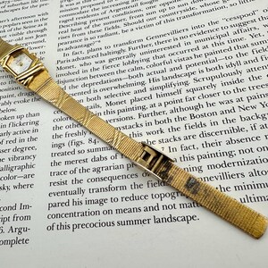 K&ouml;nnte beinhalten: Goldfarbene Armbanduhr mit rechteckigem Zifferblatt und strukturiertem Armband. Die Uhr hat ein kleines, quadratisches Zifferblatt mit goldenem Rahmen. Das Armband hat ein gewebtes Muster und einen Verschluss.