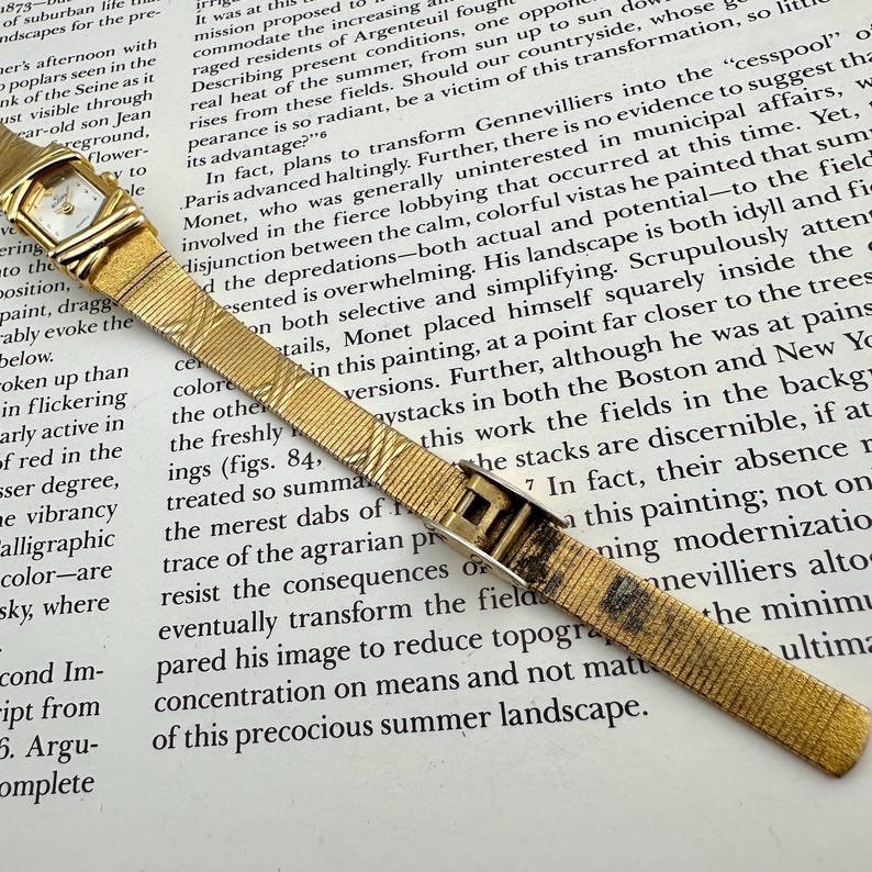 K&ouml;nnte beinhalten: Goldfarbene Armbanduhr mit rechteckigem Zifferblatt und strukturiertem Armband. Die Uhr hat ein kleines, quadratisches Zifferblatt mit goldenem Rahmen. Das Armband hat ein gewebtes Muster und einen Verschluss.
